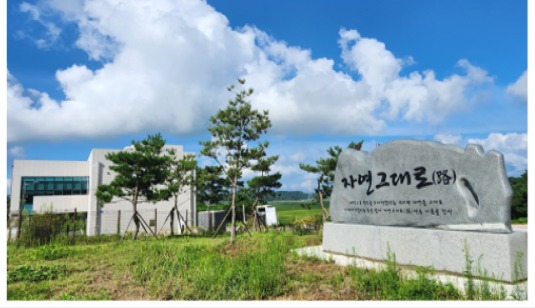 단지 입구에 세워진 각자석. 화강암에 '자연그대로(路)'가 새겨져 있고 뒤로 축사와 하늘이 보인다.